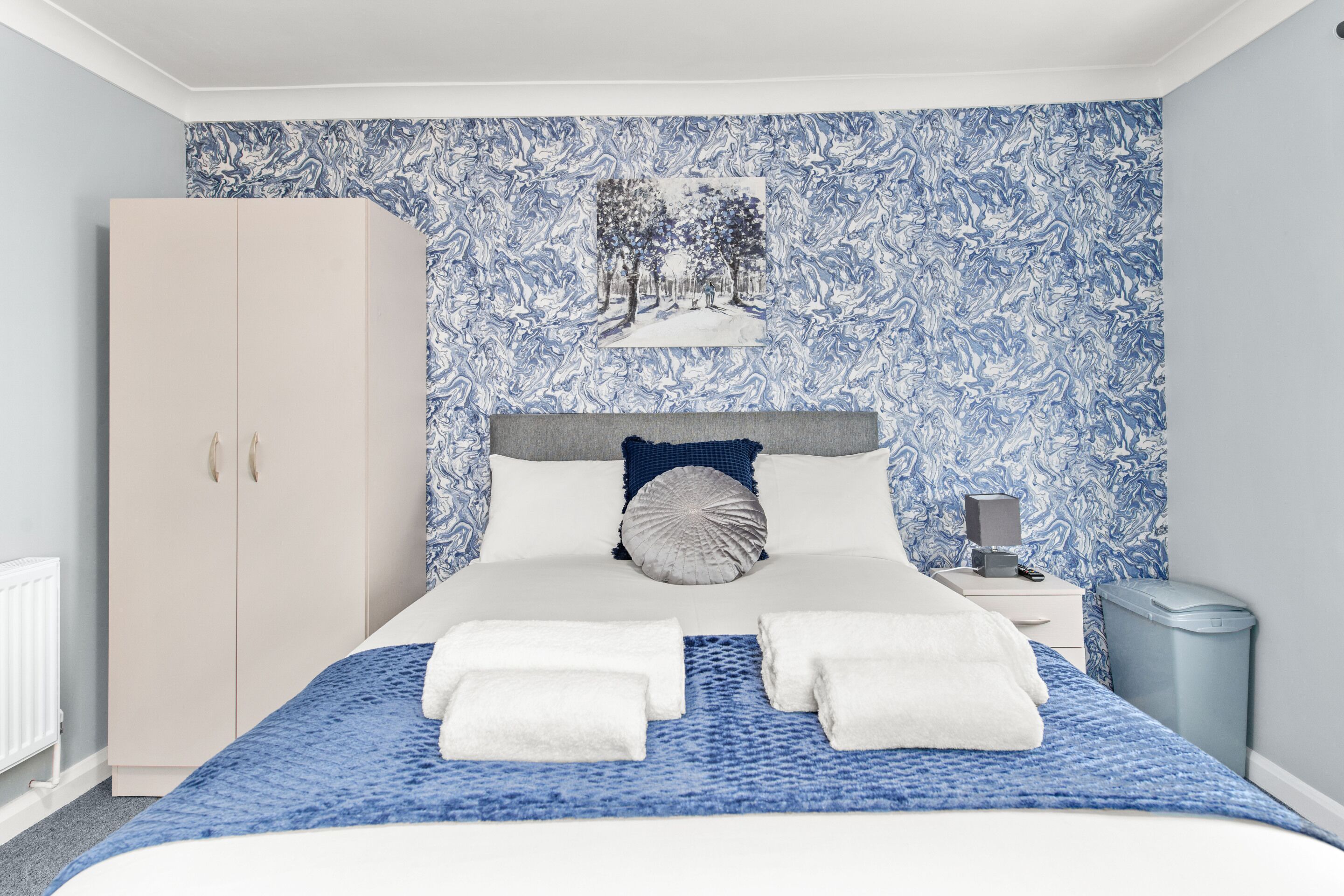 A neatly arranged bedroom with a bed, a wardrobe, a nightstand, and a trash bin.