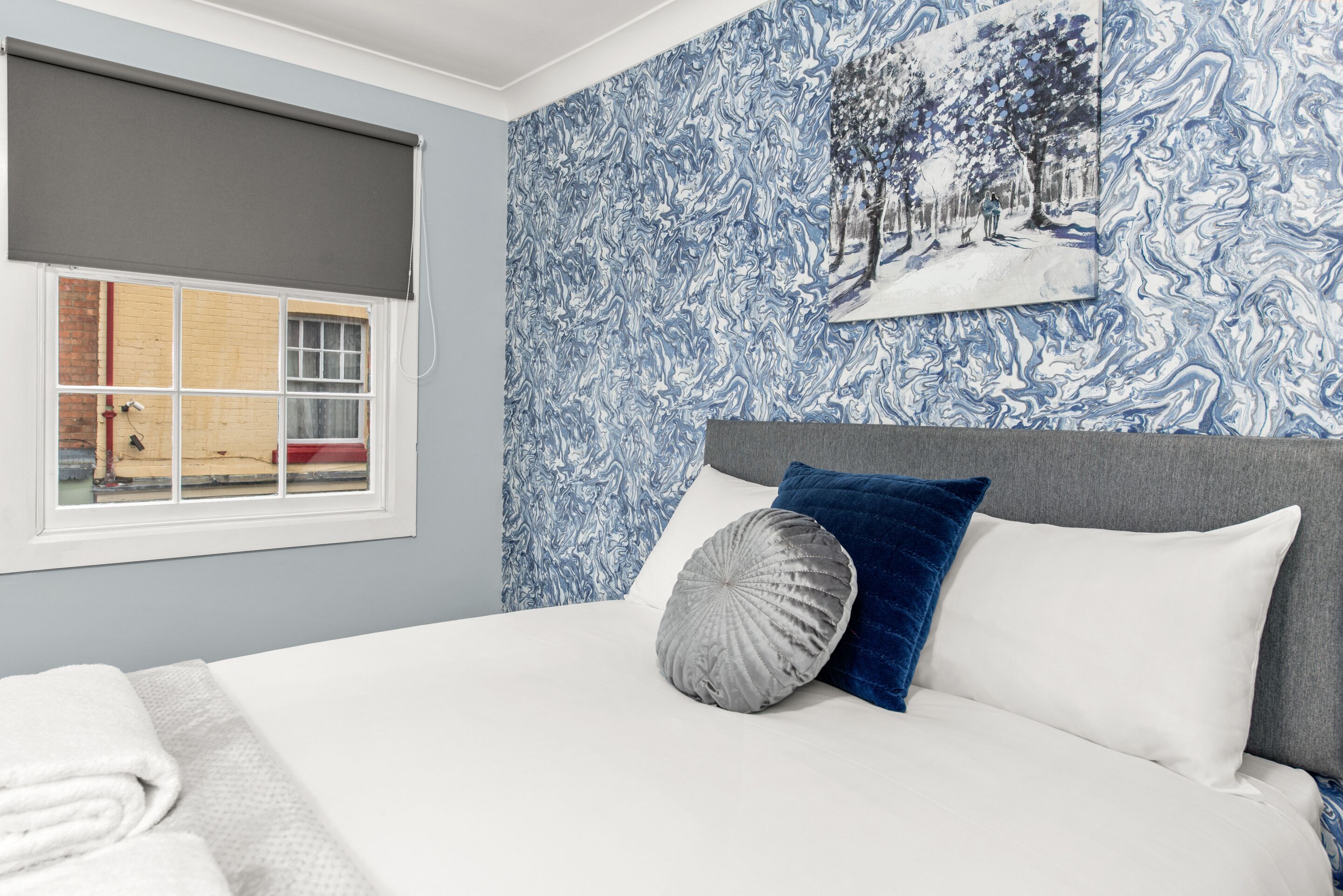 Un dormitorio con cama, ventana que ofrece vistas a un edificio y pared decorada con un diseño azul y blanco.