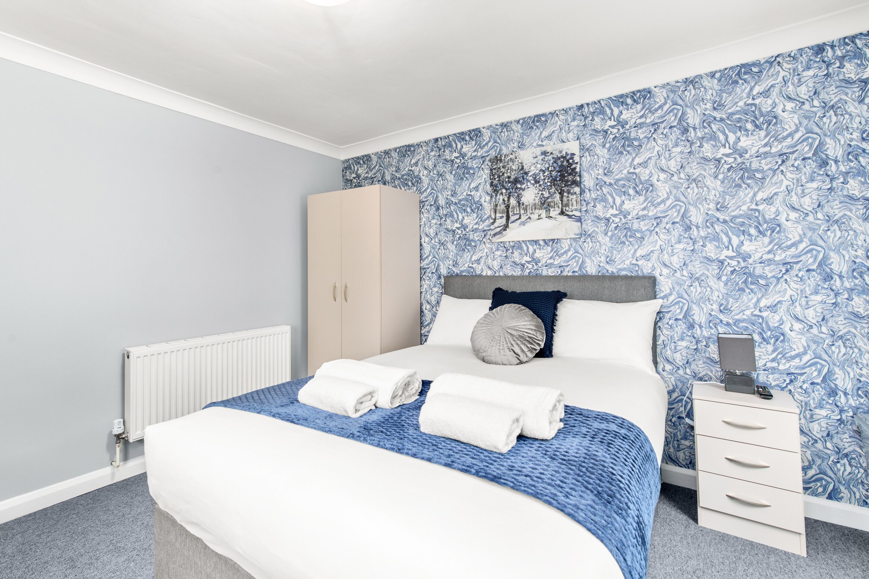 Una cama bien hecha con ropa de cama blanca y azul, una mesita de noche blanca con una lámpara y un armario.