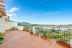 Un balcon orné de carreaux de terre cuite, de plantes en pot, et offrant une vue sur un quartier résidentiel avec des maisons et des montagnes à l’horizon.