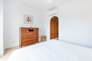 Un dormitorio con cama, cómoda de madera, cesta de mimbre, aire acondicionado de pared y puerta con arco.