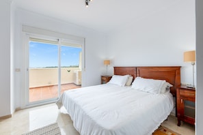 Un dormitorio con una cama grande, una mesita de noche, una lámpara y un ventanal corredera que da a un balcón.