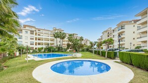 A pool area with two pools, surrounded by well-maintained grass and palm trees, with a multi-story building in the background. - Fantastic 4 bed apt La Cala Hills (Mijas Costa)