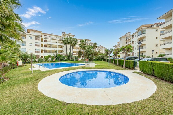 Un área de piscina con dos piscinas, rodeada de césped cuidado y palmeras, con un edificio de varios pisos al fondo.