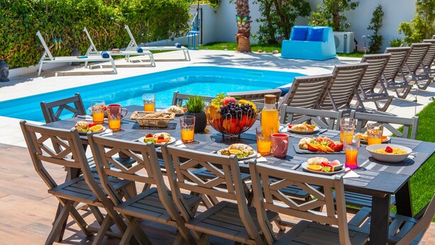 Un área de comedor junto a la piscina con una mesa dispuesta para el desayuno, rodeada de sillones de descanso y una piscina.