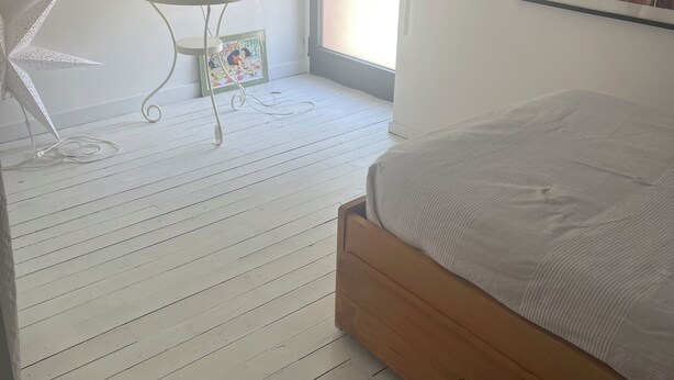 A bedroom with a wooden bed, a small table with a lamp, a chair, and a window with a view. - Chambres Dans Maison Neuve (Charly)
