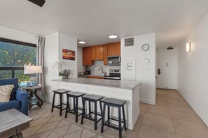 A modern kitchen with a central island, bar stools, a dining area with a sofa, and a view of greenery outside.