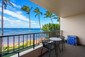 A balcony with a view of the ocean, palm trees, and a table with chairs.