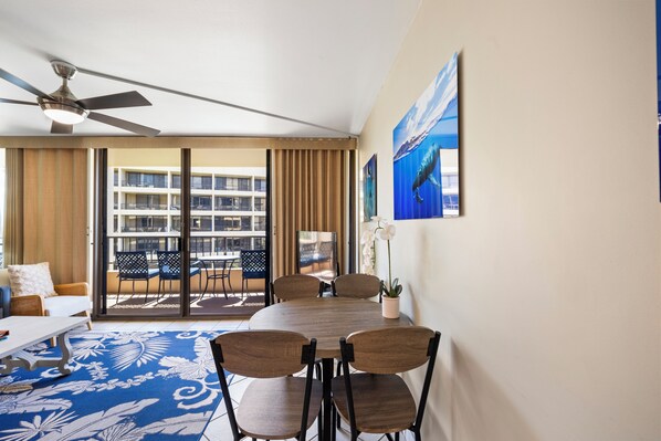 Een hotelkamer met balkon, een eettafel met stoelen, een plafondventilator en een groot raam met gordijnen.