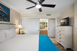 Hotelkamer met een bed, een plafondventilator, een televisie, een dressoir en een badkamer zichtbaar door een open deur.