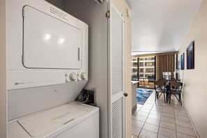 Un espace buanderie équipé d’un lave-linge et d’un sèche-linge à chargement frontal, une porte donnant sur un balcon avec vue sur les immeubles, et un coin repas avec une table et des chaises.