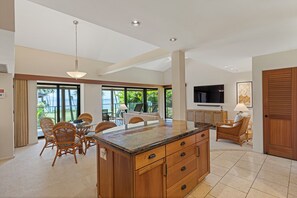 A spacious kitchen with a large island, dining area with wicker chairs, and a TV mounted on the wall. - Stunning Oceanfront 1BR/2BA Retreat with Spectacular Views - Wailea Elua 1802 (Wailea, Maui)
