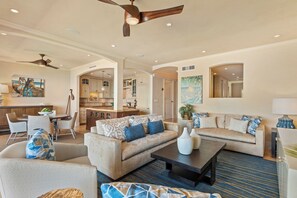 A spacious living area with a sofa, coffee table, and chairs, a dining table, and a kitchen in the background. - Luxurious Beachfront Condo with Modern Amenities - Makena Surf Resort #E-203 (Makena, Maui)