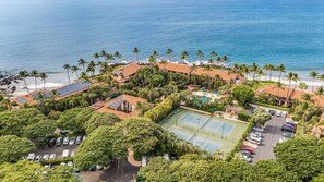 Eine Luftaufnahme eines Resorts mit Strand, Palmen, Solaranlagen auf den Gebäuden, einem Tennisplatz und einem Parkplatz.
