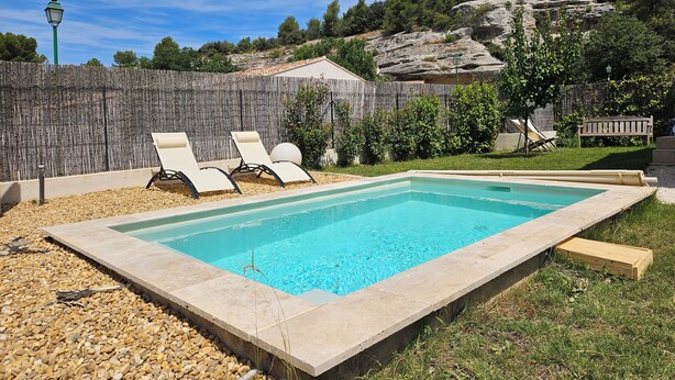 A rectangular swimming pool with a stone edge, surrounded by pebbles and grass. There are two lounge chairs and a wooden bench. A wooden fence and trees are visible in the background. - Villa des Baumes (Beaumettes)