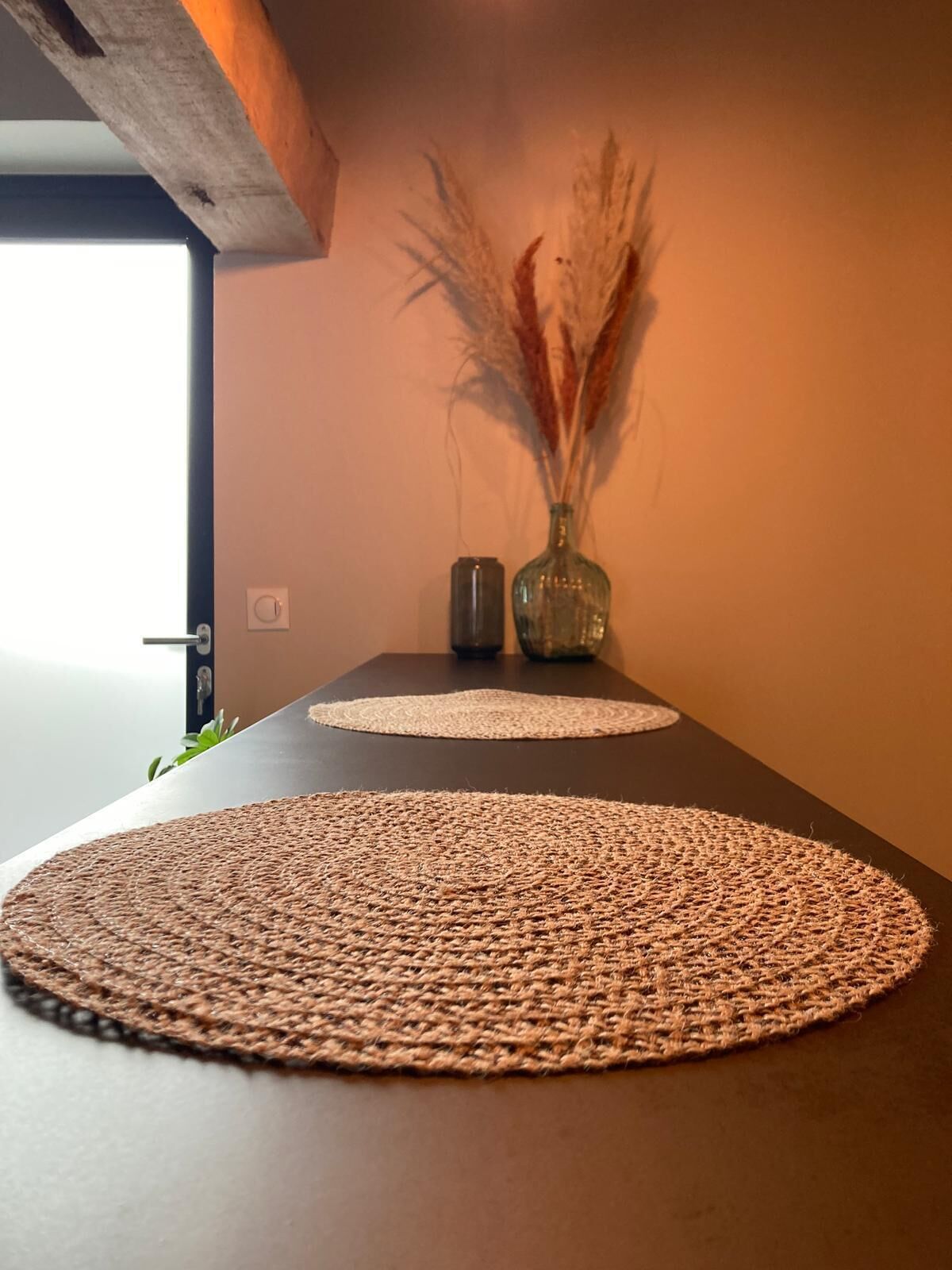 A modern room with a dark wooden table, two woven placemats, and a vase with dried pampas grass.