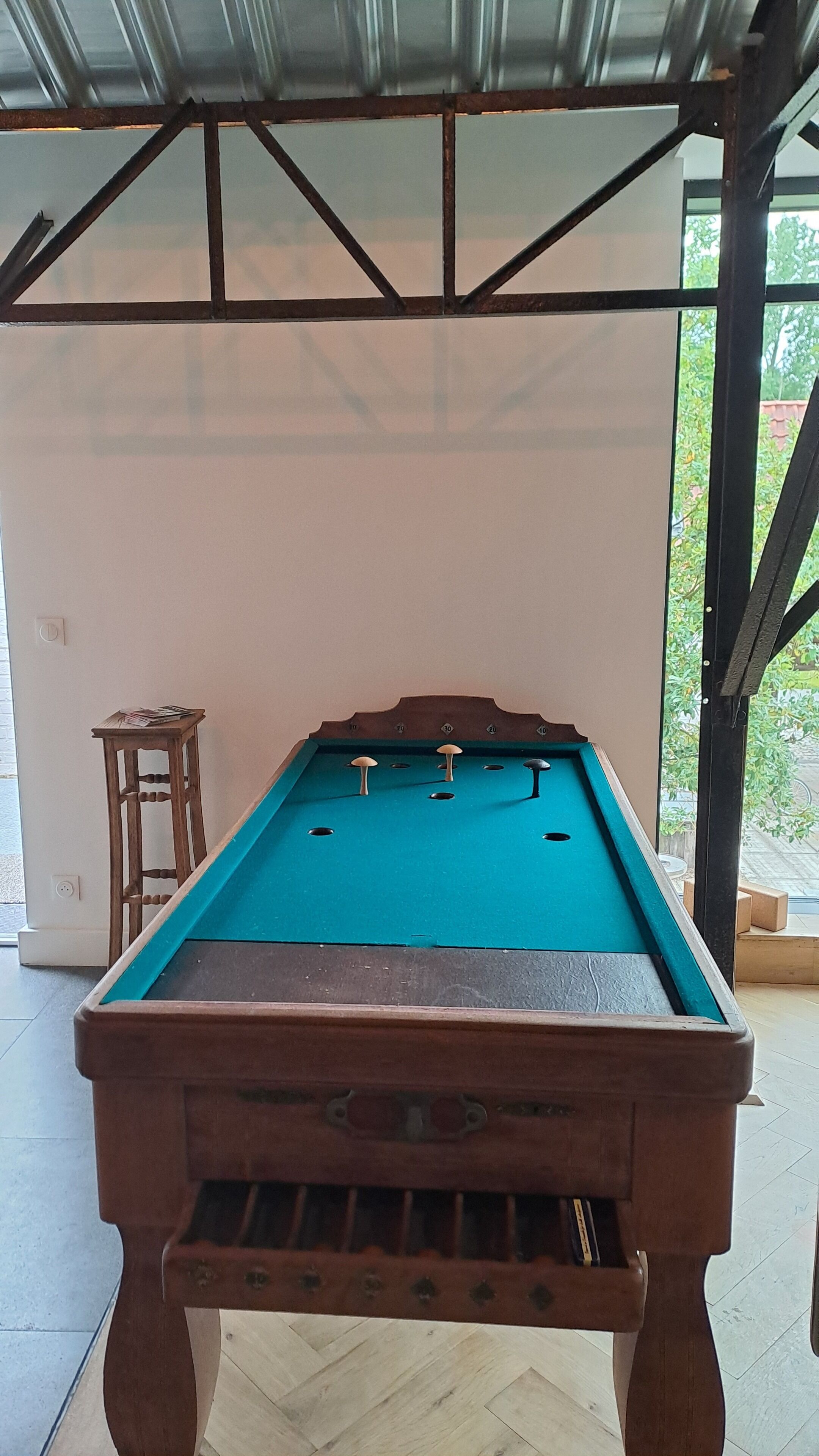 Een biljart met houten frame en een groene bekleding, in een ruimte met een metalen dak en uitzicht op groen buiten.