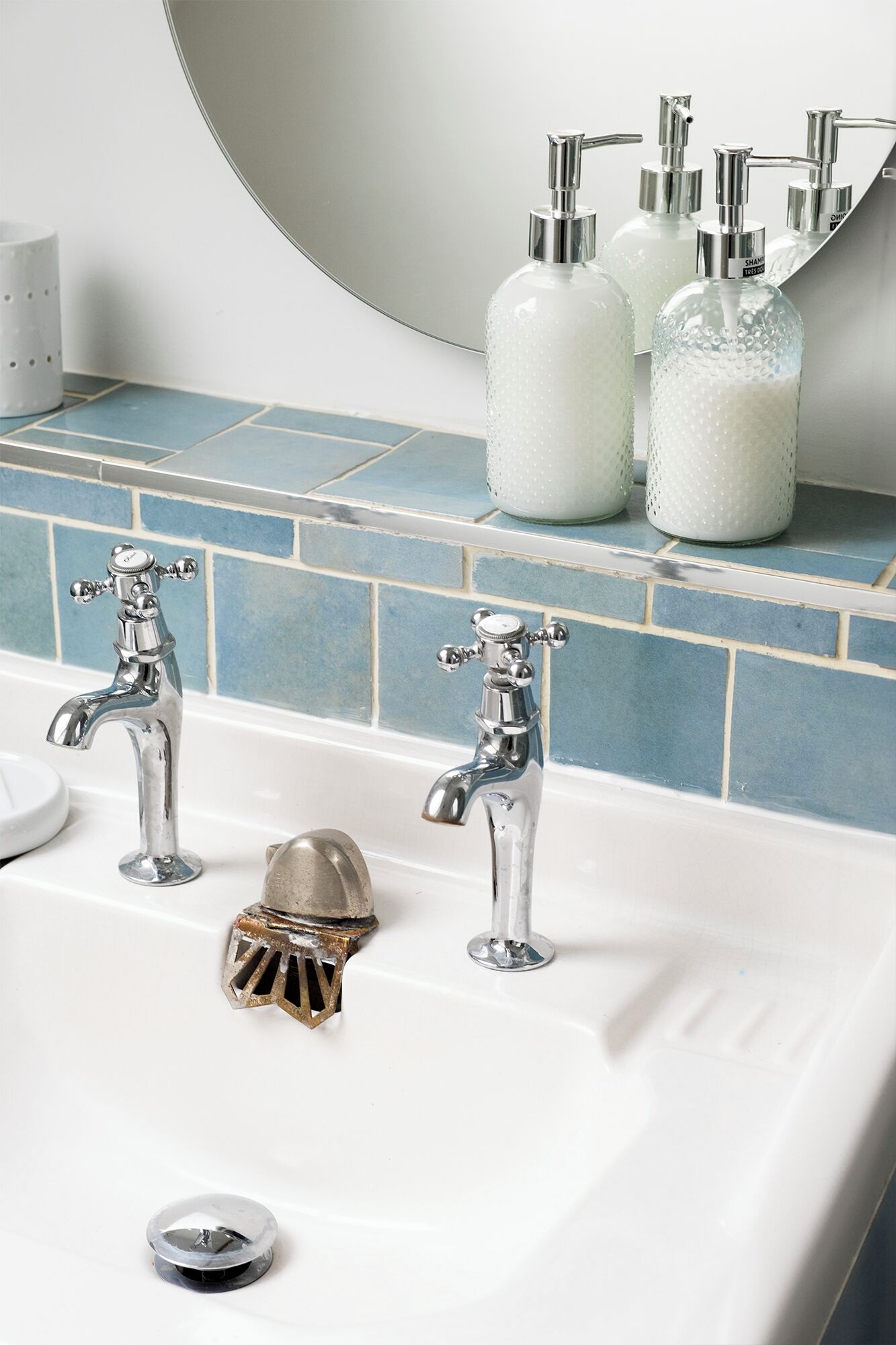 A bathroom with two chrome faucets, a white sink, and a mirror above. There are three soap dispensers on the wall.