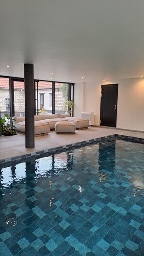 Une piscine intérieure moderne, avec un motif géométrique en mosaïque, des sièges rembourrés de couleur beige et de grandes fenêtres offrant une vue sur un bâtiment en briques.