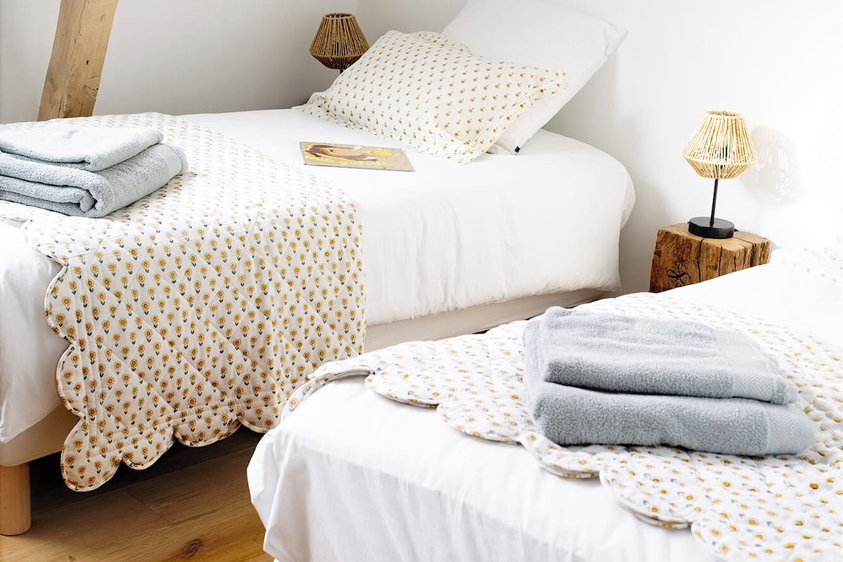 A bedroom with two beds, each with a white duvet and patterned blankets. There are two bedside tables with lamps, and the room has wooden beams on the ceiling.