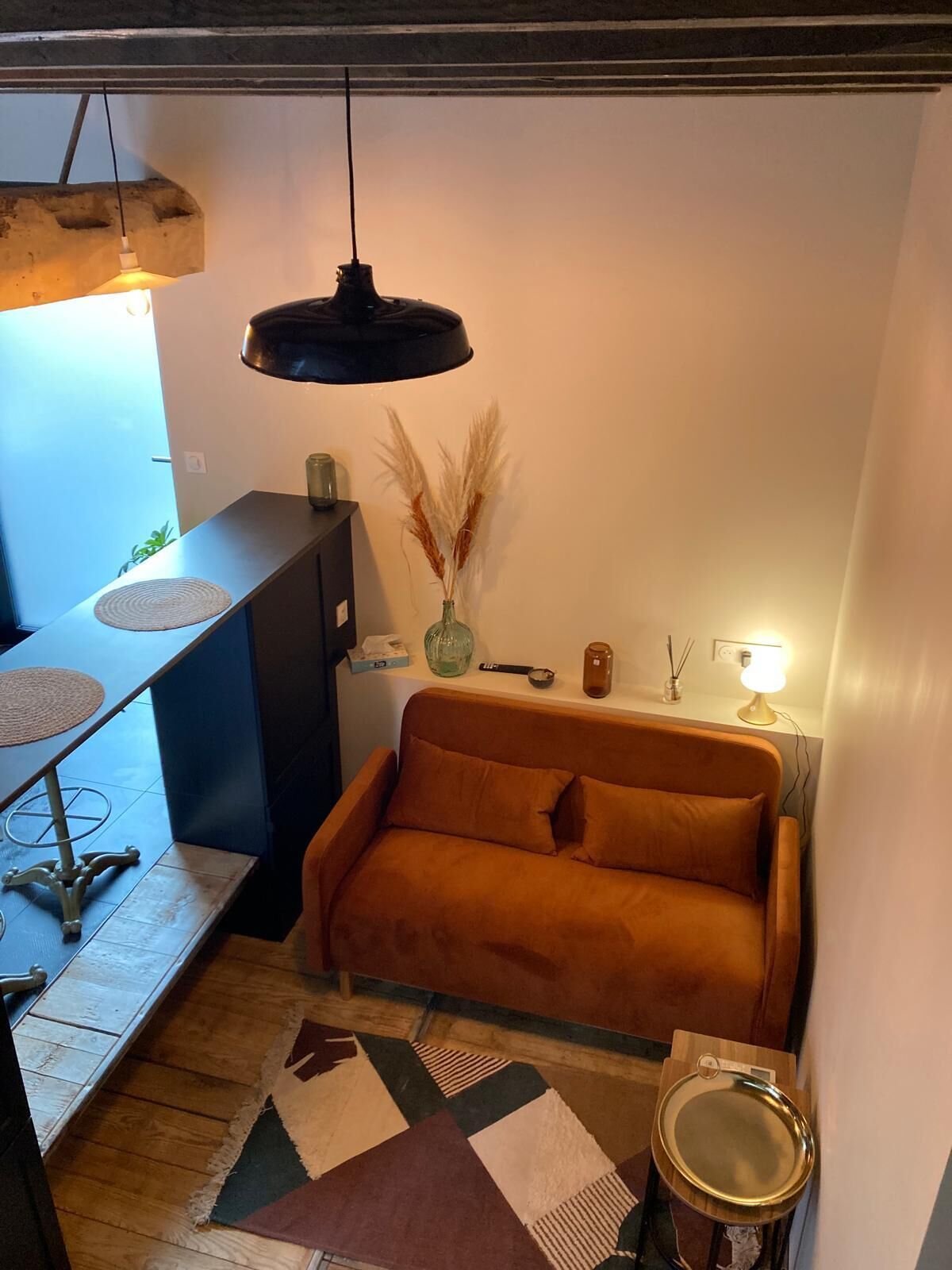 A compact living space with a sofa, a small table, a hanging light fixture, and a decorative vase with pampas grass.