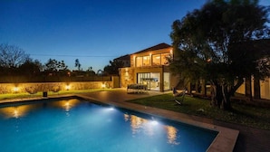 Una piscina con zona de patio, una casa de dos pisos con balcón y un espacio exterior bien iluminado.