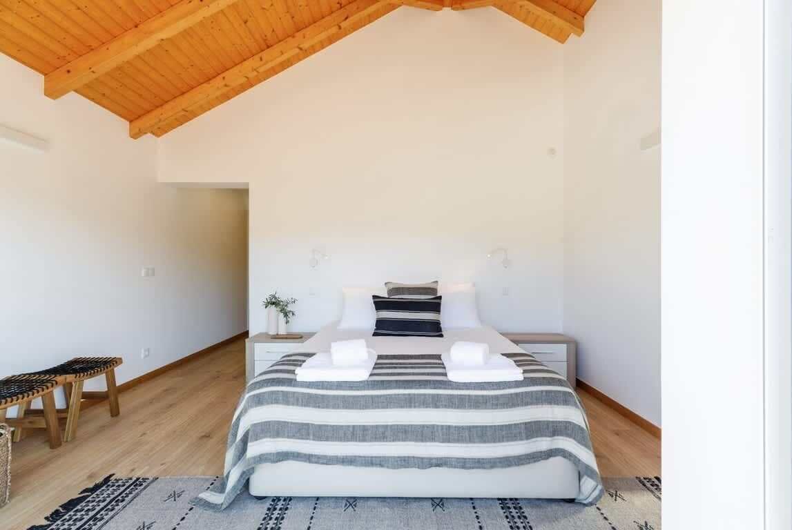 A bedroom with a wooden ceiling, a bed with striped bedding, a bedside table, a bench, and a rug.