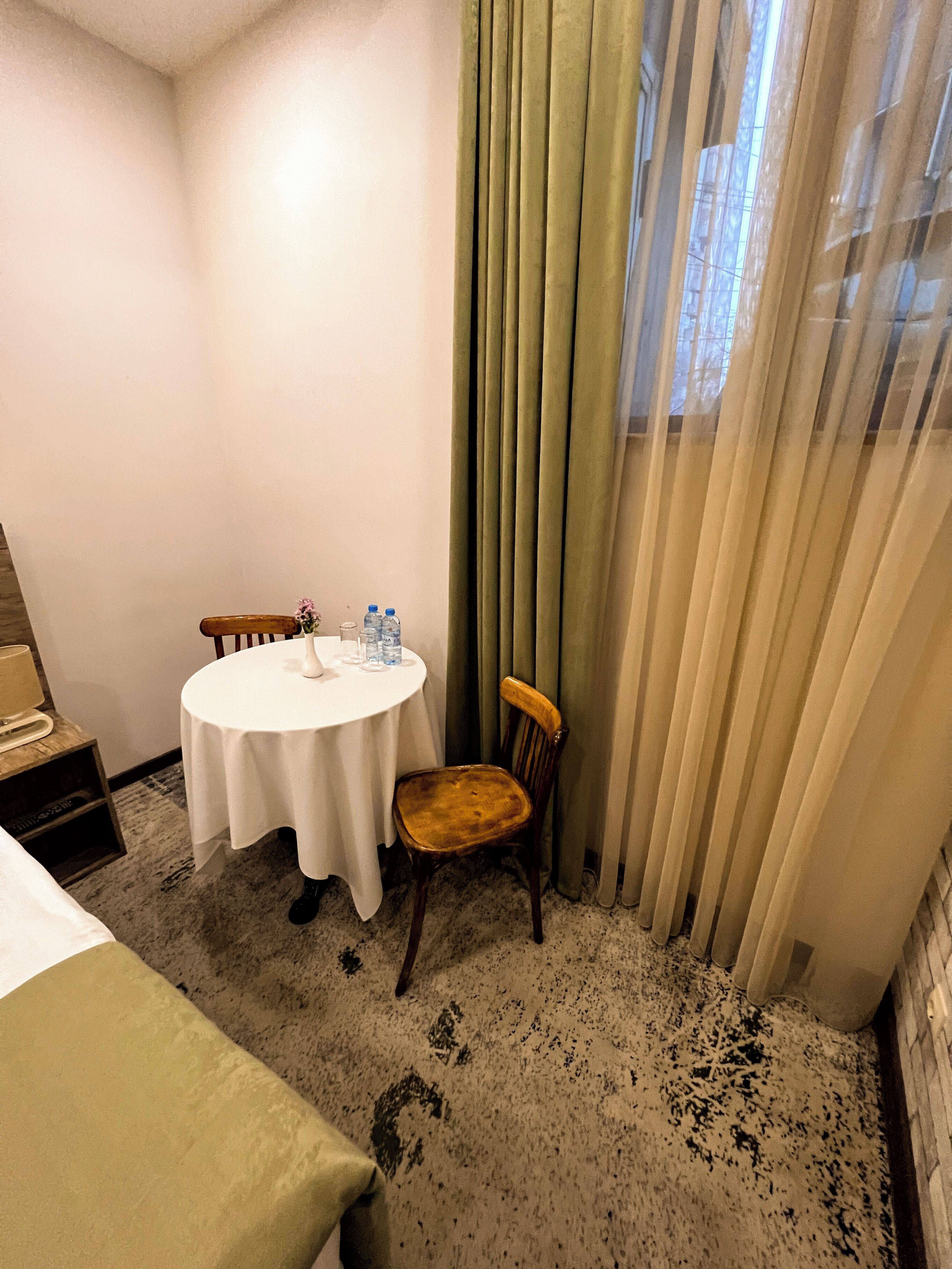 Hotellihuone, jossa on pyöreä pöytä, kaksi tuolia, sänky, yöpöytä, lamppu ja ikkuna, jossa on verhot.