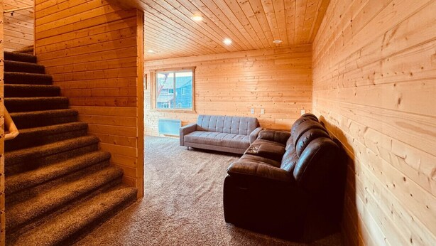 Interior de una cabaña de troncos con una escalera, un sofá gris y un sofá de cuero.