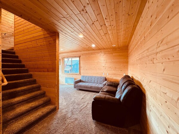 Interior de una cabaña de troncos con una escalera, un sofá gris y un sofá de cuero.