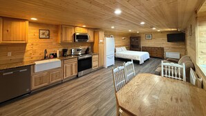 Interior acogedor de una cabaña con paredes y pisos de madera, una cocina con electrodomésticos modernos, un comedor y un dormitorio con cama y cómoda.