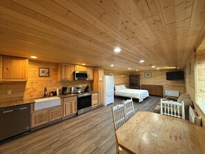 A cozy cabin interior with wooden walls and floors, a kitchen with modern appliances, a dining area, and a bedroom with a bed and dresser. - 12 miles from Yellowstone with incredible views - Grizzly Lodge (Island Park)