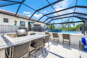 Eine Terrasss mit Grill, Sitzgelegenheiten und Blick auf einen Swimmingpool.
