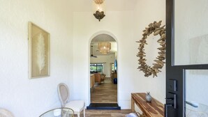 Een hal met een witte muur, een lijst met een veer kunstwerk, een rieten stoel, een glazen tafel, een houten bank met een ananas decoratie en een houten wanddecoratie.