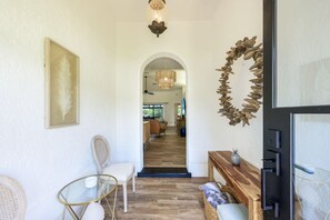 Ein Flur mit weißer Wand, einem Bilderrahmen mit Federkunstwerk, einem Korbstuhl, einem Glastisch, einer Holzbank mit Ananasdekor und einer Wanddekoration aus Holz.