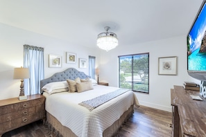 A bedroom with a large bed, two bedside tables, a chandelier, a TV, and a window with blinds. - Resort Like Amenities Close to Beach (Bonita Springs)