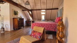 A cozy attic bedroom with a wooden ceiling, a bed with a floral bedspread, a wicker chair, and a dresser. - Luxe Appartement in het Oude Centrum (Zutphen)
