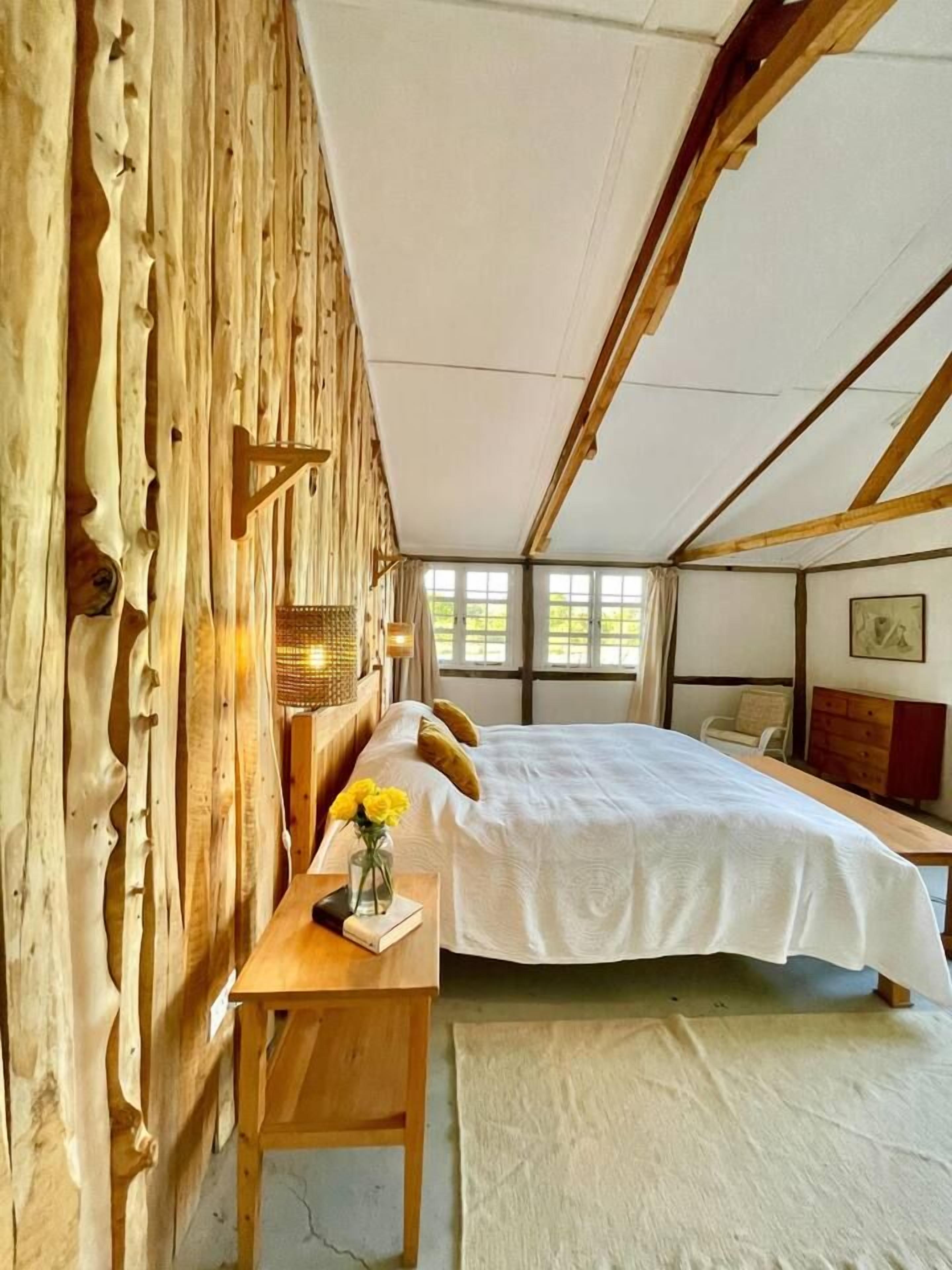 A room with a bed, a wooden desk, a vase with flowers, and a window.