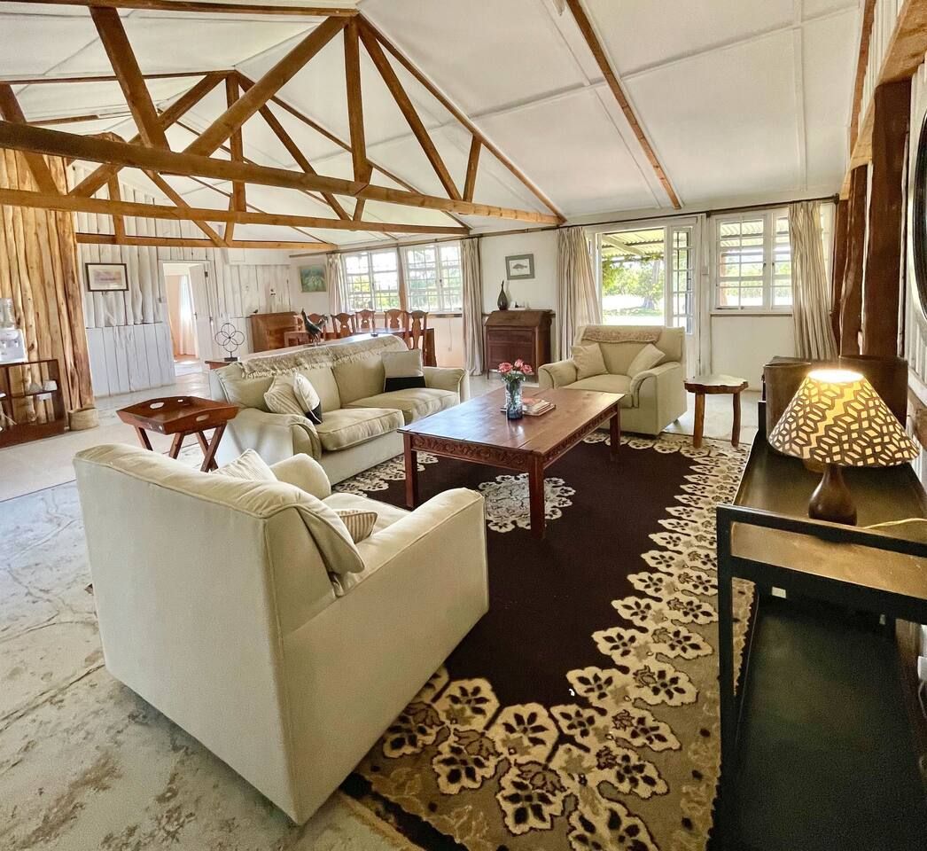 A spacious living room with a high ceiling, wooden beams, and large windows. There are multiple seating areas with sofas and a coffee table.