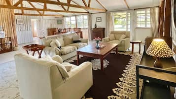 A spacious living room with a high ceiling, wooden beams, and large windows. There are multiple seating areas with sofas and a coffee table.