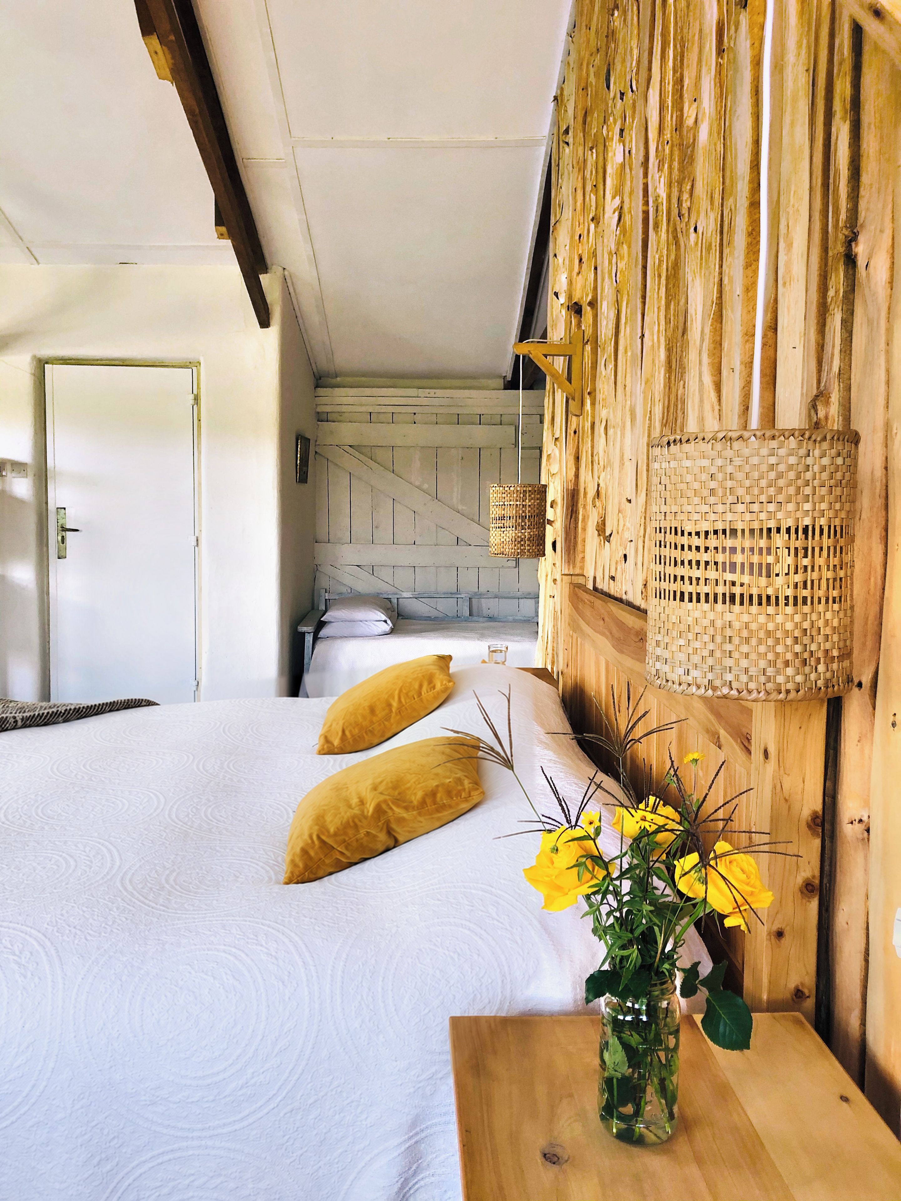 A room with a bed, a wooden wall, a woven lamp, and a vase with flowers.