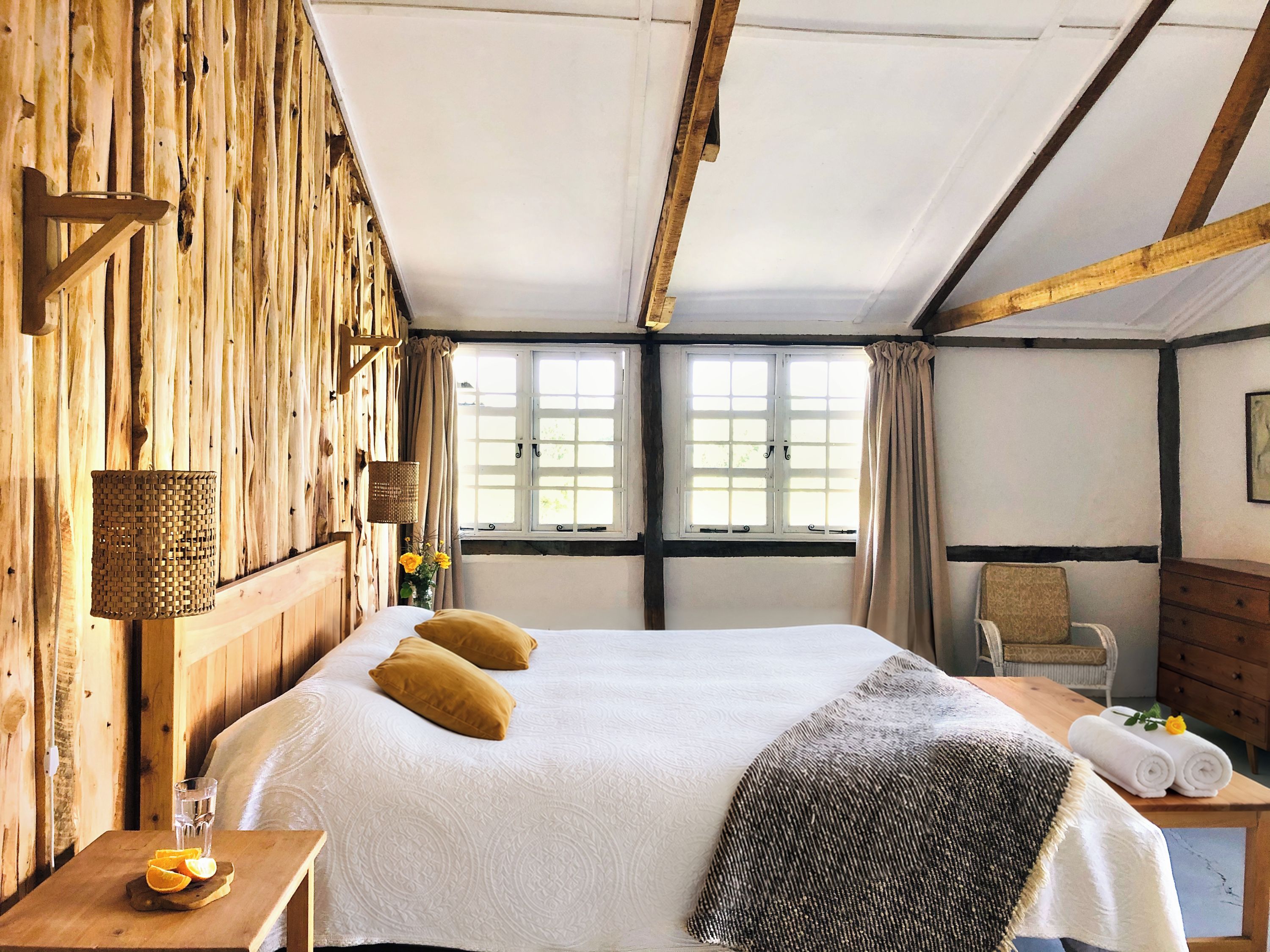 A wooden room with a bed, a table with a glass of water and oranges, and a window with curtains.