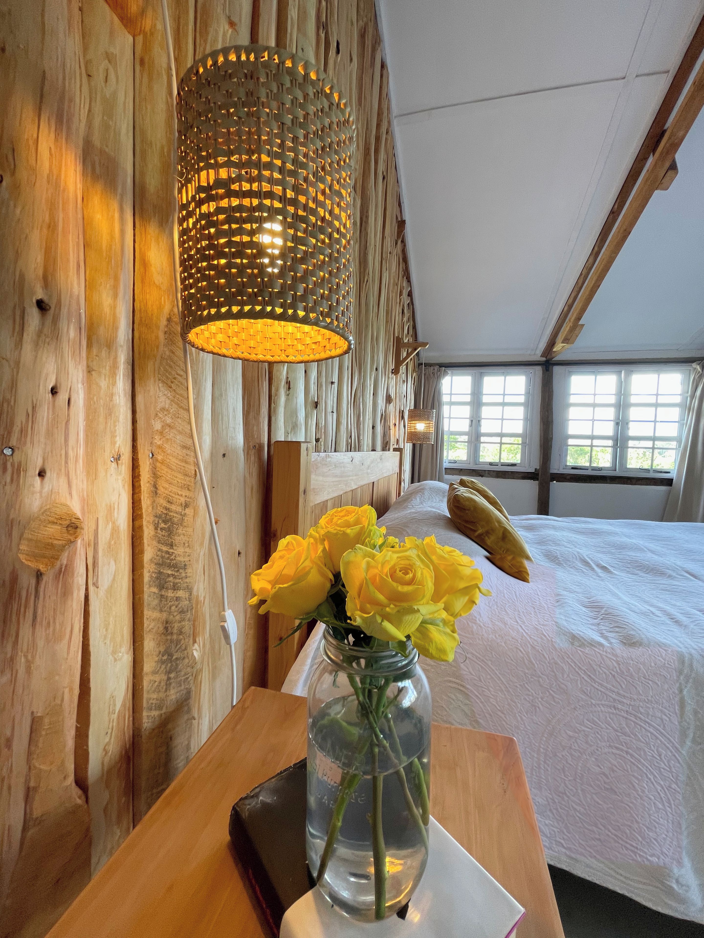 A wooden interior with a bed, a vase with yellow flowers, and a hanging light fixture.
