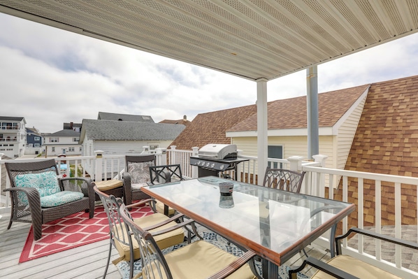 A covered outdoor deck with a glass table and chairs, a grill, and a view of neighboring houses.