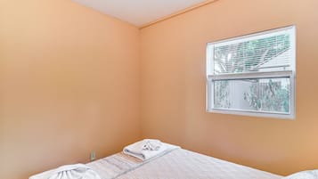 A single bed with a quilted bedspread and a folded towel, a window with blinds, and a wall with a power outlet.