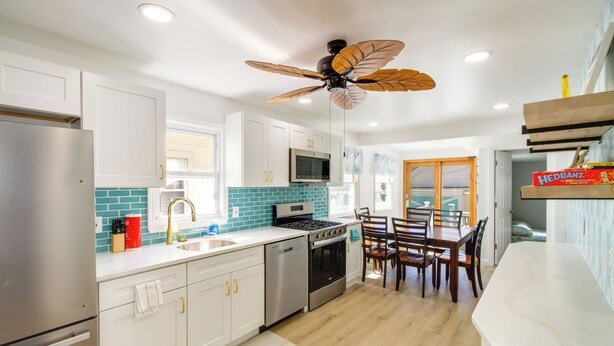 A modern kitchen with white cabinets, a stainless steel refrigerator, a microwave, and a stove. There is a ceiling fan with wooden blades, a dining area with wooden chairs, and a kitchen island with a sink. - Wildwood Group Retreat: 2 Blocks to Boardwalk! (North Wildwood)