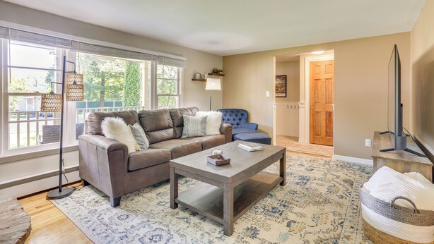 A living room with a brown leather sofa, a coffee table, and a blue armchair. There is a large window with blinds, a floor lamp, and a television on a stand. - Charming Highland Falls Home < 2 Mi to West Point! (Highland Falls)