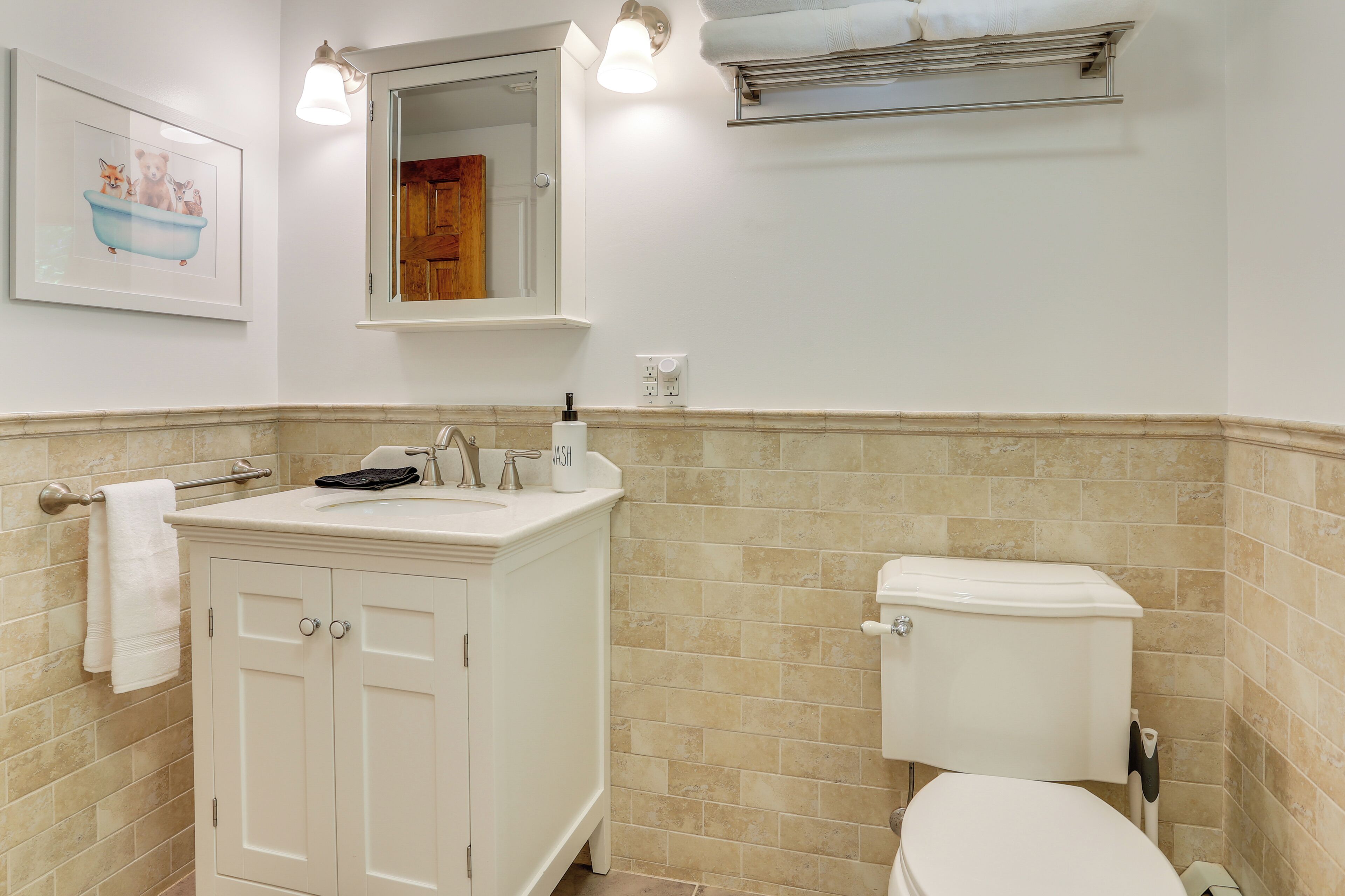 Casa de banho com lavatório branco, espelho e sanita. Tem um porta-toalhas, um quadro emoldurado na parede e um candeeiro sobre o espelho.