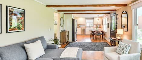 A modern living room with a gray sofa, a beige armchair, and a coffee table. There is a kitchen in the background with wooden beams on the ceiling.