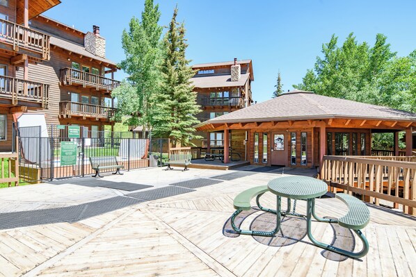 A wooden deck with picnic tables and benches, surrounded by buildings with balconies and a covered seating area. - Streamside! Ski In/Out & Hiking (Breckenridge)